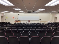 Front of room view with lectern on left in front of markerboard and exit doors on left and right