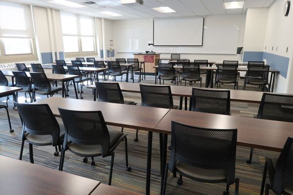 Front of room view with lectern on left in front of markerboard and projection screen lowered