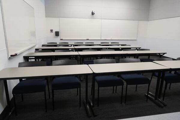 Back of room view of student table and chair seating, camera on rear wall, and markerboards on rear and right side wall