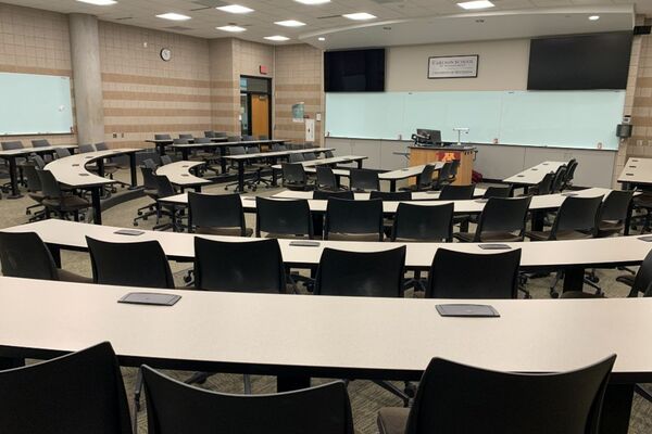 Front of room view with lectern center in front of markerboard, exit door on front left, chairs and fixed tables 