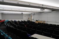 Front of room view with lectern on left of demonstration bench in front of markerboard, exit door on left side of front wall