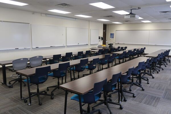 Front of room view with lectern on right in front of markerboards, additional markerboards on right side wall of room, and door located in right corner of room