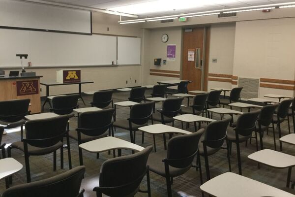 Front of room view with lectern on left in front of markerboard, projection screen partially raised, and exit door on right wall