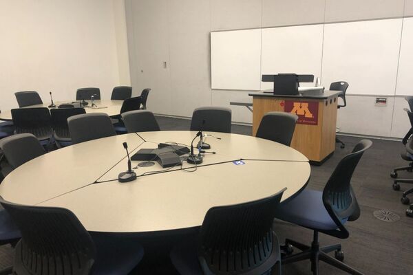 View of student collaborative table and chair seating, lectern at center right of room in front of markerboard