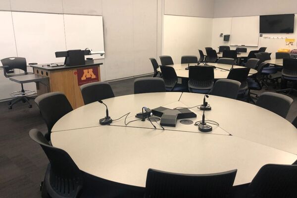 View of student collaborative table and chair seating, lectern towards the left in front of markerboard, monitor and markerboard on right wall