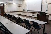 Front of room view with lectern on right in front of markerboard and projection screen lowered