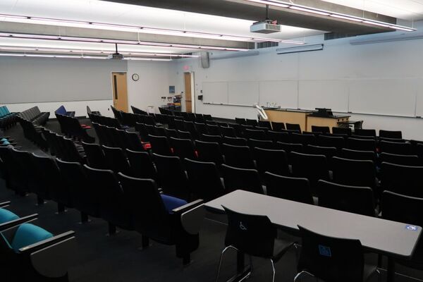 Front of room view with lectern on right side of demonstration bench in front of markerboard, exit door on left side of front wall