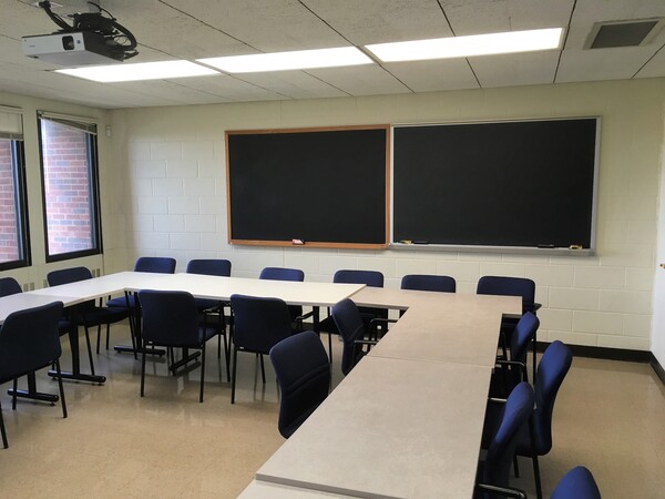 Photo of back of room from front of room, tables arranged in a U shape, windows on left wall.