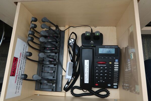 Pedestal - inside view of drawer showing two wireless mics in charging base, telephone, and assistive listening devices in charger