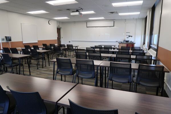 Front of room view with lectern on right in front of markerboard, projection screen partially raised, and exit at left front of room