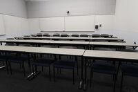 Back of room view of student table and chair seating, camera on back wall, markerboards on back and left side wall