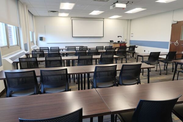 Front of room view with lectern on right in front of markerboard and projection screen lowered, exit door on right wall