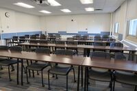 Back of room view of student table and chair seating and, camera and three markerboards on back wall of room