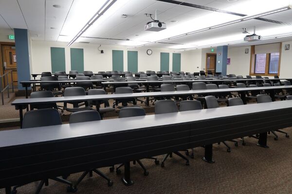 Back of room view of student tiered fixed-table and chair seating and exit doors at left and right rear of room