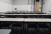 Back of room view of student table and chair seating, markerboards on left and rear wall, and exit door at right rear of room