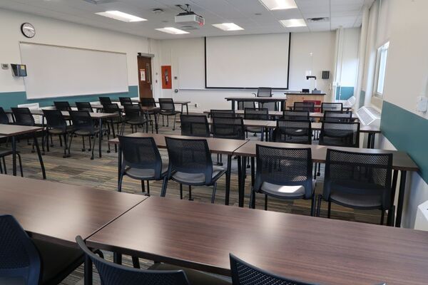 Front of room view with lectern on right in front of markerboard and projection screen lowered, exit door in left corner of room