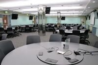 Room view with lectern in center, markerboards on all walls, exit door center left wall, and projection screen on right wall