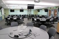 Room view of student active learning table seating, markerboards on all walls, and two exit doors at right side of room