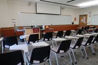 Front of room view with lectern on left in front of markerboard, exit door to the right, and projection screen partially raised