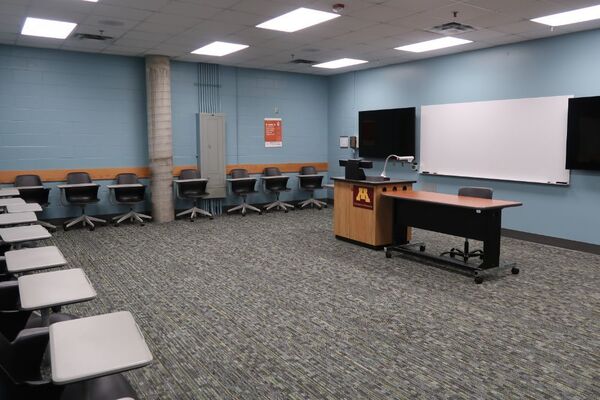 Front of room view with lectern center in front of markerboard and display screens to the left and right, exit door on the right