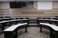 Back of room view of student tiered fixed table and chair seating and dual confidence monitors to left and projection screen to right