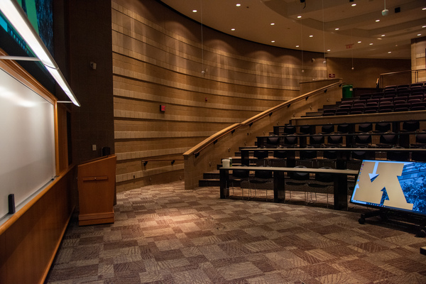 Instructor view from front right of room looking towards whiteboard/projectors.  Illuminated whiteboard, projector display, and handicap entrance/exit are visible.  