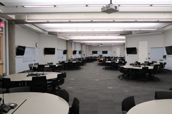 Room view with lectern in center of room, walls of room have multiple markerboards and student display monitors, three exit doors on left wall of room