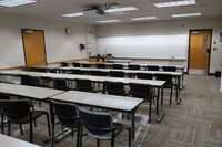 Front of room view with lectern on left in front of markerboard, exit door to the right, table and chair seating