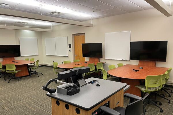 View of student table and chair seating, student table displays, markerboards, and exit door on the right