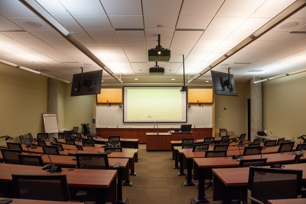 View from audience of center of classroom.  Projector and front TVs are visible, along with exit door on right and student seating and microphones.  Room camera is also visible.