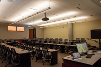 View facing audience from right side perspective showing backside of instructor podium, projector, rear exit door, instructor camera, rear self view monitors, and audience seating