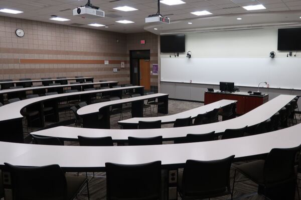 Front of room view with lectern on center in front of markerboard and display screens to left and right, exit door on the left