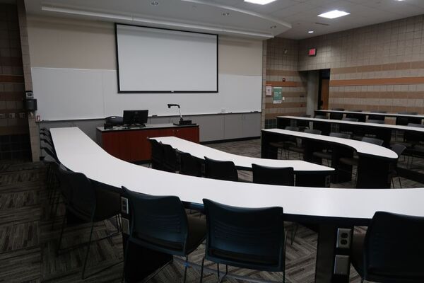 Front of room view with lectern on in front of markerboard and projection lowered, with exit door on the right