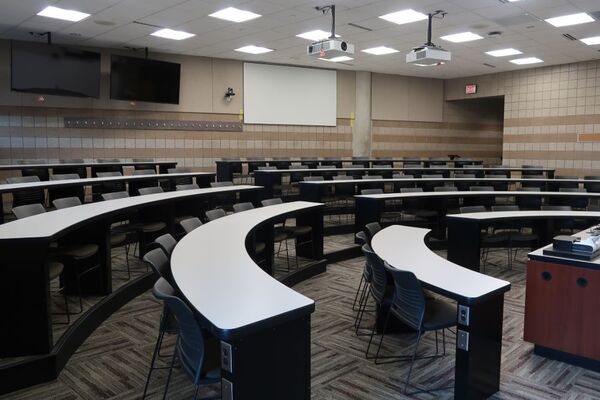 Back of room view of student tiered fixed-table and chair seating, project screen and confidence monitor on rear wall, exit door at right rear of room