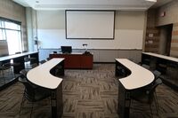 Front of room view with lectern on left in front of markerboard and projection screen lowered with exit door to the right